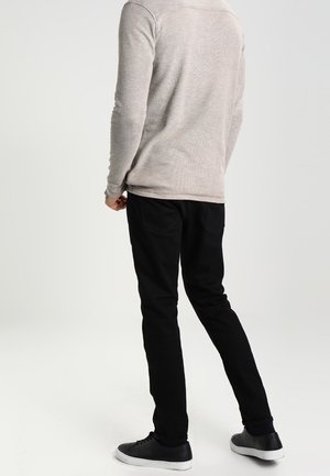 Adult wearing a light gray long-sleeve shirt, black pants, and black sneakers with white soles, standing facing away on a white background.