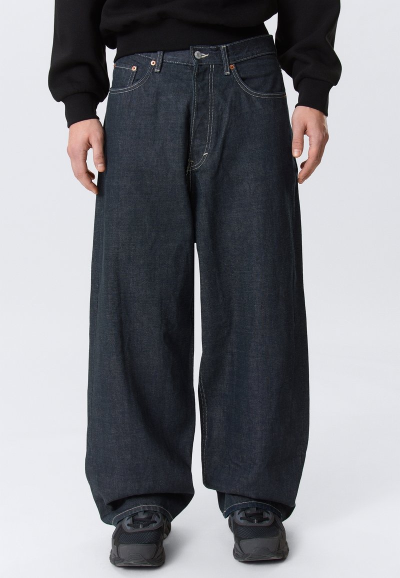Personne portant un jean en denim foncé à jambes larges, des baskets noires et un haut noir à manches longues, debout devant un fond uni.
