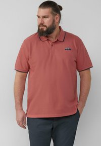 Mann mit Bart und Dutt, der ein rostfarbenes Poloshirt mit Auto-Stickerei und dunklen Hosen trägt und nach links schaut, während eine Hand in der Tasche ist.