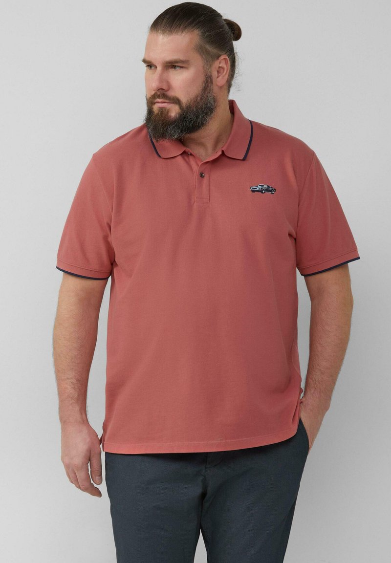 Mann mit Bart und Dutt, der ein rostfarbenes Poloshirt mit Auto-Stickerei und dunklen Hosen trägt und nach links schaut, während eine Hand in der Tasche ist.