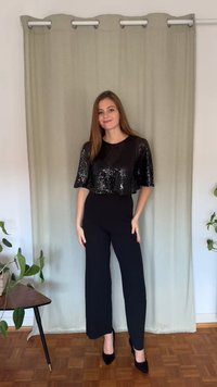 Melns spīguļots īss tops, kopā ar melniem, vidukļa augstuma biksēm ar platiem galiem. Modele valkā melnus stiletto papēžus, stāvot pret gaišu aizkaru.