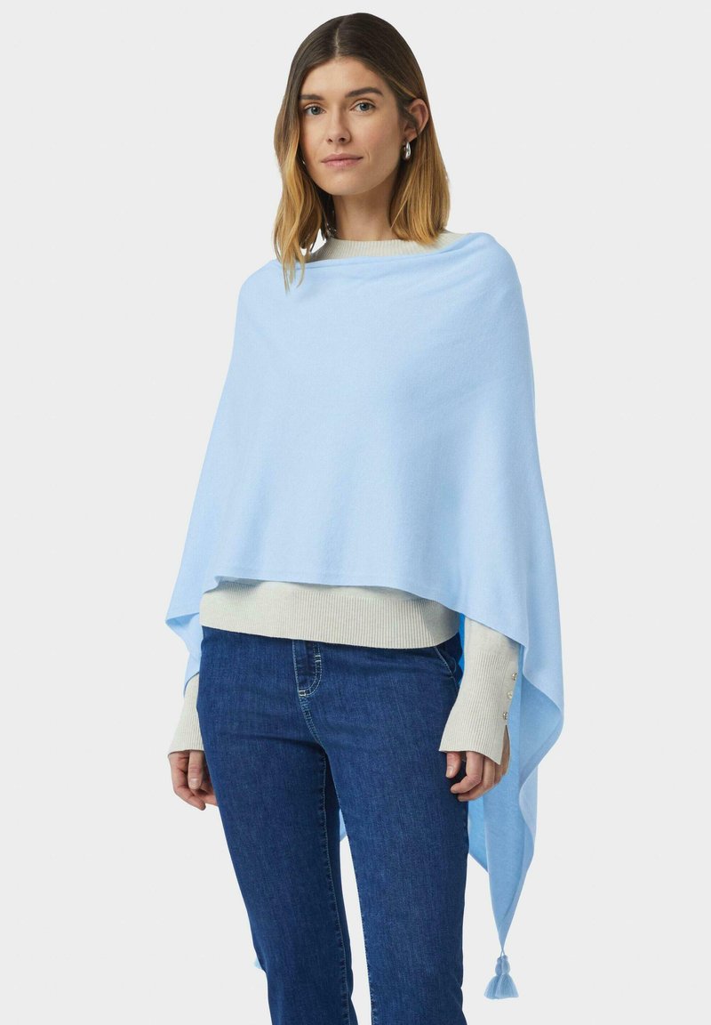 Kobieta w jasnoniebieskim szalu z frędzlami, nosząca beżowy sweter i niebieskie dżinsy, stojąca na tle białej ściany.