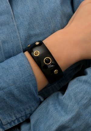 Bracelet jonc noir brillant avec des accents en métal doré et des détails circulaires. Porté au poignet, posé sur un fond en denim.