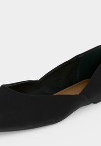 Μαύρο μαλακό δερμάτινο παπούτσι μπαλαρίνα με μυτερή μύτη, χαμηλό τακούνι και καφέ επενδυμένο πάτο, σε λευκό φόντο.