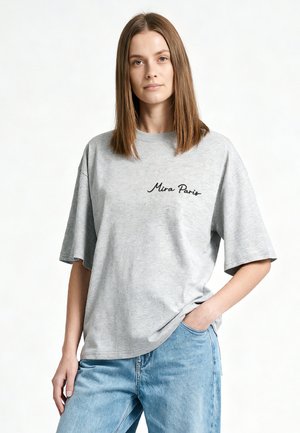 Jonge vrouw met stijlbruin haar, een los grijs T-shirt met de tekst 'Mira Paris' en lichtblauwe spijkerbroek, staand met één hand in de broekzak.
