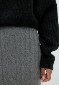 Schwarzer gestrickter Pullover mit langen Ärmeln über einem grauen Zopfmuster-Rock. Der Stoff hat eine strukturierte Optik, die einen geschichteten Eindruck erzeugt.