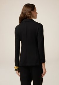 Blazer nero su misura con colletto a punta, maniche lunghe e polsi arrotolati, caratterizzato da una texture liscia e una silhouette strutturata.