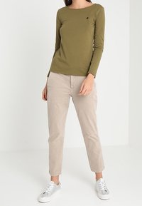 Olivgrünes Langarmoberteil mit rundem Halsausschnitt, kombiniert mit hellbeigen Hosen. Das Modell trägt silberne Sneakers. Einfaches Design, keine Muster.