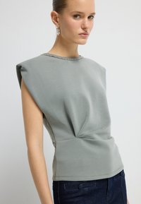 Giovane donna con top grigio senza maniche dotato di spalline imbottite e jeans blu scuro, in piedi su uno sfondo bianco semplice.