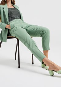 Mintgröna skräddarsydda byxor med en slät yta, ihop med en randig topp. Modellen bär gröna blockklackar medan hon sitter.