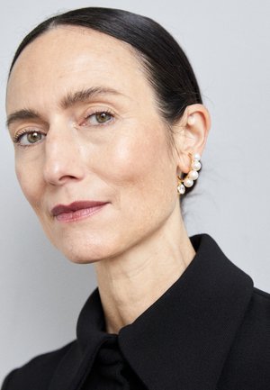 Zlati uhani v obliki obročka z biseri in kristalnim detajlom, ki jih nosi oseba s gladko kožo in temnimi lasmi, oblečena v črno oblačilo s ovratnikom.