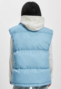 Svetlomodrá pufovaná vesta s hladkým, lesklým povrchom, bez rukávov a viditeľným horizontálnym prešívaním, je prehodená cez biely hoodie.