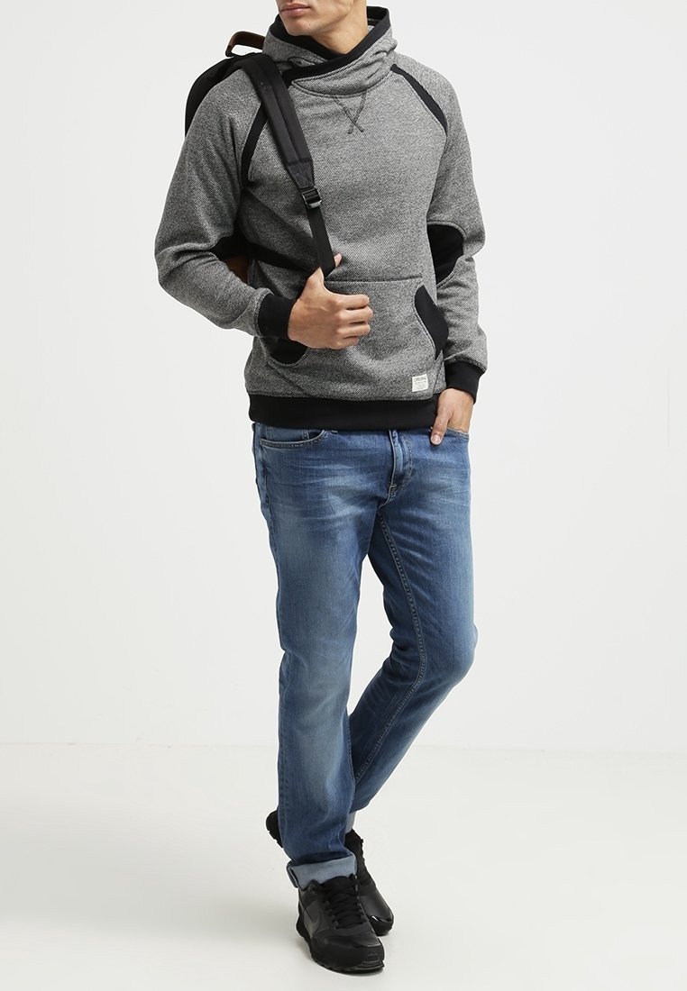Sudadera con capucha en tonos gris y negro, con un alto cuello, bolsillo frontal y mangas raglán, combinada con unos vaqueros azules y zapatillas negras.