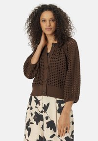 Cardigan en crochet marron avec un col rond, fermeture à boutons et manches trois-quarts. Motif de tissage ouvert remarquable et ourlet côtelé.