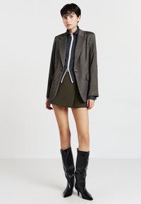Una donna con un blazer marrone sopra un top scuro con zip, una mini gonna verde oliva e stivali neri lucidi fino al ginocchio, in piedi davanti a uno sfondo bianco.