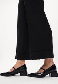 Melna ādas Mary Jane papēžu kurpes ar smailu purngalu, potītes siksniņu un bloku papēdi. Valkātas ar melnām platām biksēm, kuru apakšdaļa ir redzama.