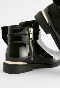 Schwarze Lackleder-Ankle Boots mit einem seitlichen Reißverschluss und einem goldenen Akzent an der Ferse. Mit einem weichen Textil-Ziehband an der Rückseite.