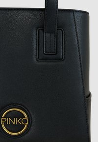 Borsa a mano in pelle nera con logo rotondo dorato, texture liscia e design strutturato con accenti metallici rettangolari sulle maniglie.