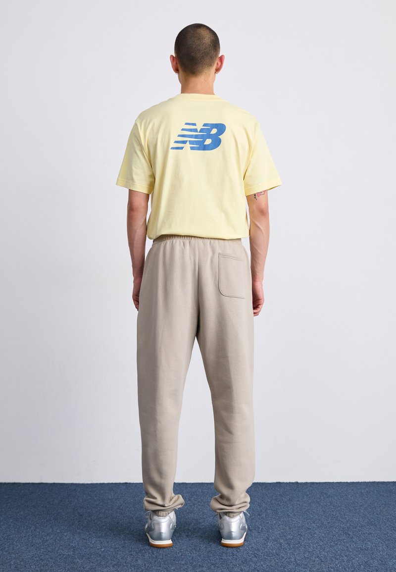 Gelbes Baumwoll-T-Shirt mit blauem "NB"-Logo auf dem Rücken, kombiniert mit hellbeigen Sweatpants, die über eine Seitentasche und eine lockere Passform verfügen.