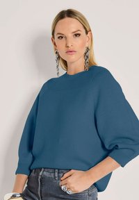 Frau mit glattem blonden Haar, die große silberne Kettenohrringe, einen blauen strukturierten Pullover mit Ballonärmeln und dunkelgraue Jeans trägt.