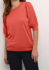 Frau trägt einen korallenfarbenen Kurzarmpullover mit gerippten Puffärmeln und schwarze, figurbetonte Hosen, steht vor einem schlichten weißen Hintergrund.