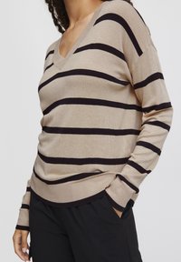 Beiger V-Ausschnitt-Pullover mit schwarzen horizontalen Streifen, gestrickte Textur, lange Ärmel und gerippte Bündchen, getragen über schwarzen Hosen.