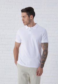 White polo shirt with a classic collar and short sleeves. Features button placket and ribbed sleeve hems. Paired with light beige trousers.