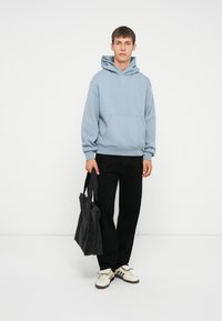 Hoodie azzurro chiaro con tasca frontale, indossato con pantaloni neri e sneakers color crema. La persona tiene una borsa tote nera.