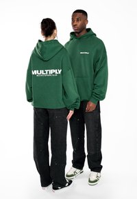 Sudaderas verdes con el texto blanco "MULTIPLY", combinadas con jeans negros holgados y zapatillas. Presenta un bolsillo tipo canguro y un ajuste oversize.