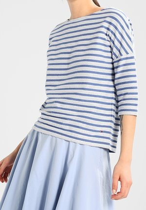 Personne portant une chemise à manches longues rayée bleu et blanc et une jupe bleu clair, montrée du cou à mi-cuisse.