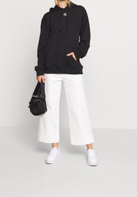 Černá mikina s kapucí, bílé široké kalhoty a bílé tenisky. Obsahuje klokaní kapsu a logo. Černá taška s krátkým popruhem.