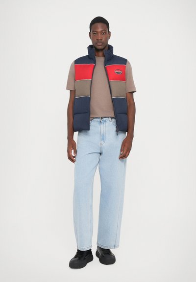 Nachtblau en rode donzen gilet met een rits, voorzien van horizontale kleurblokken en een logo-patch, gedragen over een bruine t-shirt en lichtblauwe jeans.