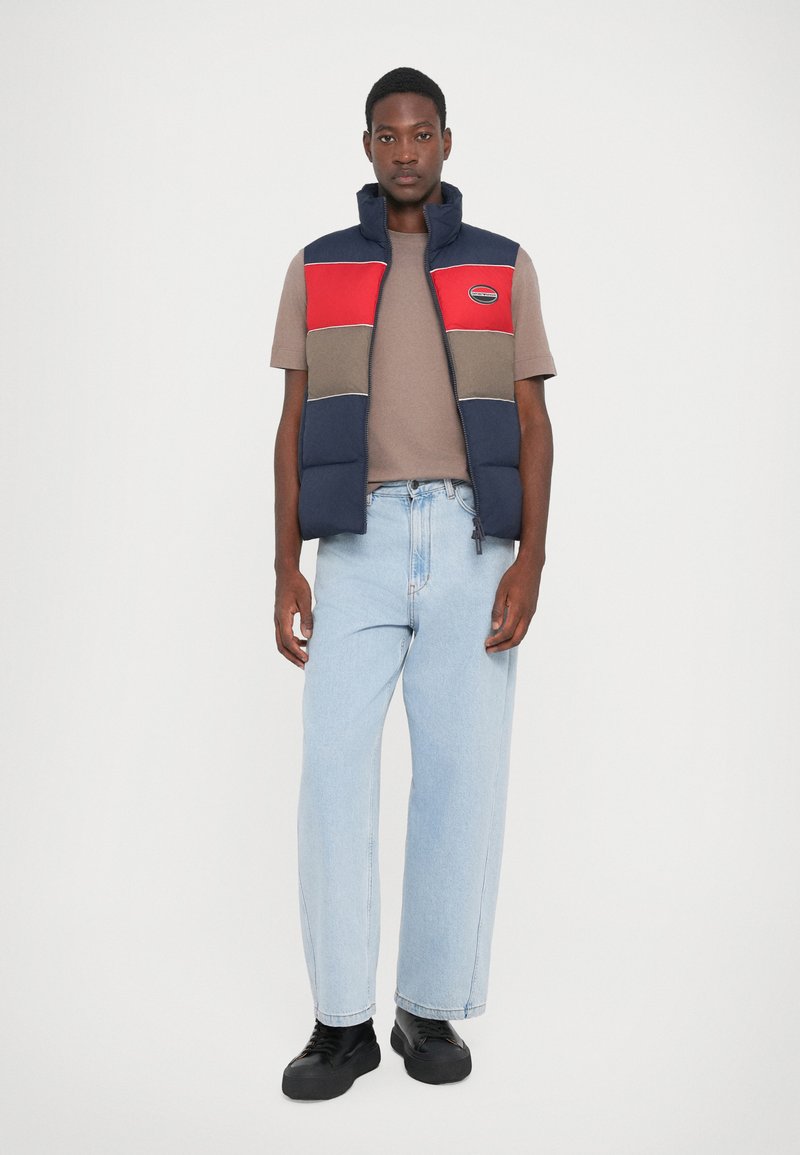 Marinblå och röd pufferväst med dragkedja, med horisontella färgblock och en logotyp-patch, deltagande över en brun t-shirt och ljusblå jeans.
