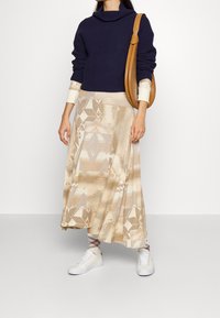 Marineblauwe gebreide trui met een col, gecombineerd met een beige, patroonrijke maxi-rok met geometrische ontwerpen. Witte sneakers en een lichtbruine handtas.