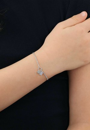Feines silbernes Kettenarmband mit zwei kleinen herzförmigen Anhängern, getragen am Handgelenk vor einem dunklen, gerippten Stoffhintergrund.