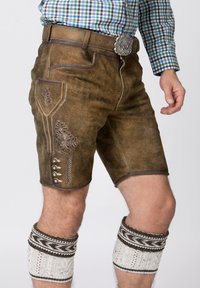 Pantaloni in pelle marrone con cuciture decorative, una tasca laterale e dettagli in bottone argentato, abbinati a gambaletti lavorati a maglia bianchi con motivo.