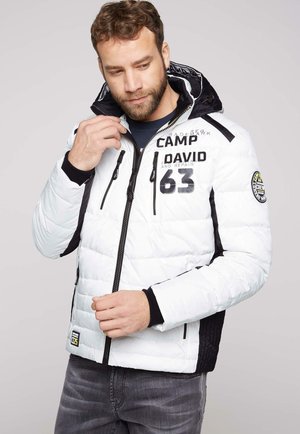 Mann passt den Kragen einer weißen gesteppte Jacke mit schwarzen Akzenten, Schriftzügen und Aufnähern an, trägt graue Jeans vor einem schlichten hellen Hintergrund.