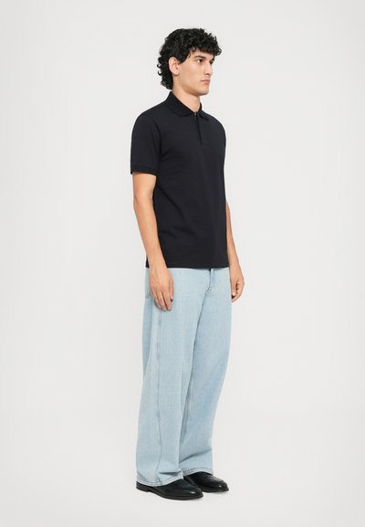 Manlig modell som står på sidan iförd en svart kortärmad polo, ljusblå vida jeans och svarta skor, mot en enfärgad bakgrund.