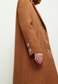 Personne portant un manteau marron texturé à double boutonnage avec des boutons argentés sur les manches et à l'avant, sur un fond clair uni.