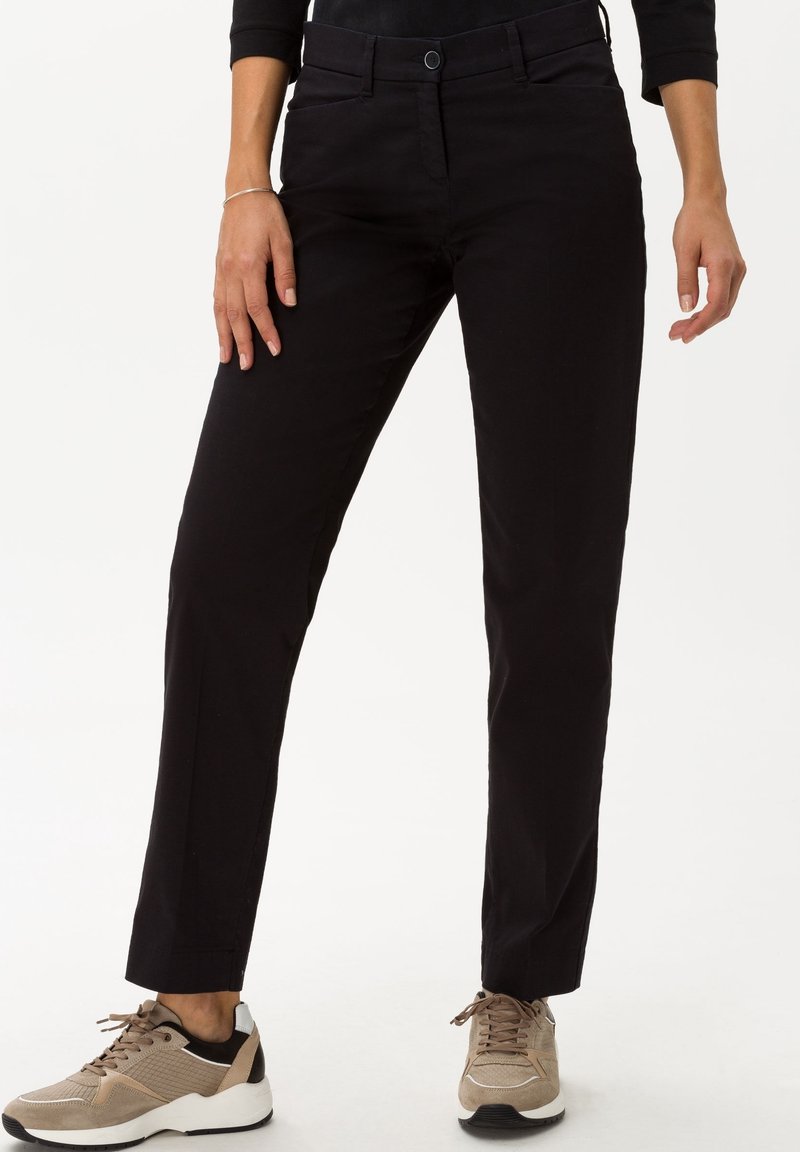 Femme portant un pantalon droit noir et des baskets beiges, debout les mains détendues le long du corps sur fond blanc.
