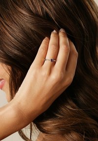 Silberring mit einem zentralen runden Edelstein in dunklen Farbtönen, ausgestattet mit einem glatten Band und minimalistischem Design. Die Hand ruht auf braunem Haar.