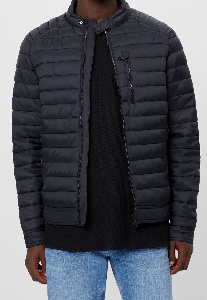 Man i en svart vadderad jacka med dragkedjefickor över en svart skjorta och ljusblå jeans, som står framför en enfärgad bakgrund.