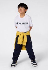 Weißes T-Shirt mit "CHAMPION"-Logo, gelbes Sweatshirt um die Taille gebunden, dunkelblaue Jogginghose und schwarze Sneaker mit gelben Akzenten.