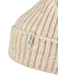 Beige knit beanie featuring a ribbed texture and a folded cuff. Includes a small tag with the brand name stitched on the side.