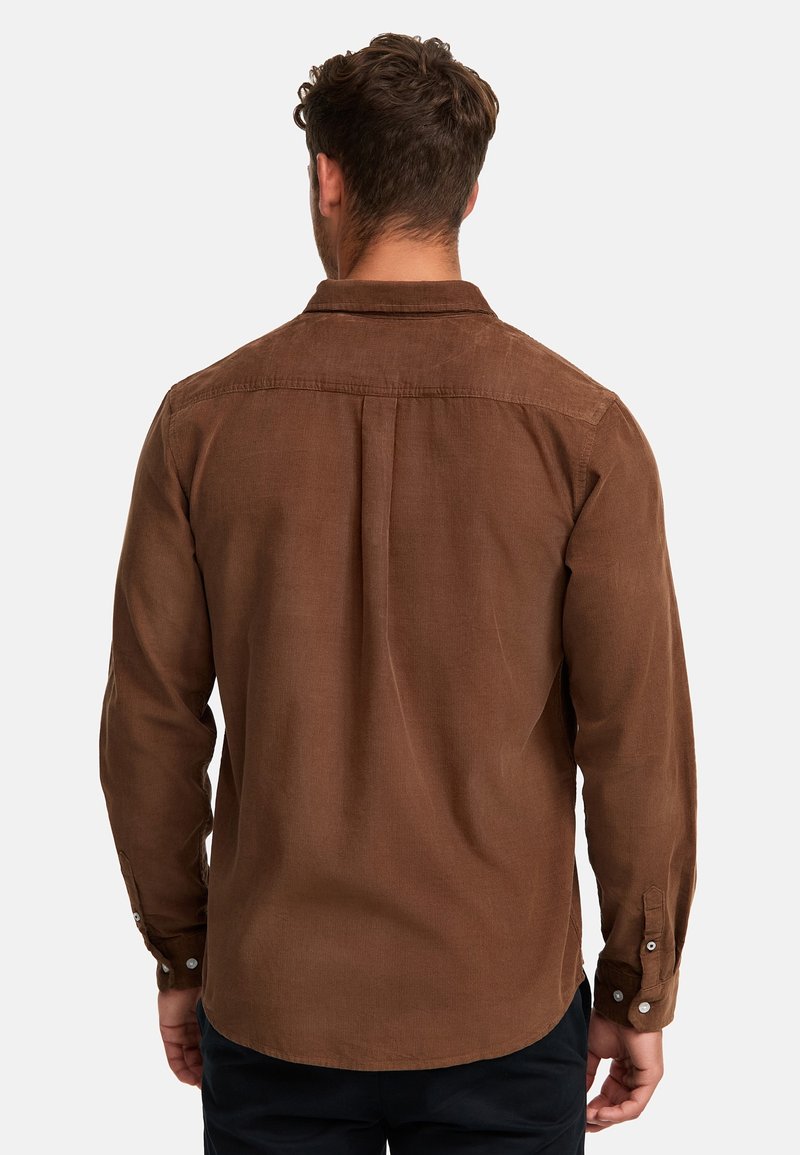 Homme portant une chemise marron à manches longues et un pantalon noir, vu de dos sur un fond uni.