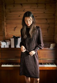 Bruine metallic jurk met lange mouwen en een getailleerde taille met ceintuur, gemaakt van gestructureerde stof met een losse pasvorm. Op de achtergrond staan een piano en cadeauverpakkingen.