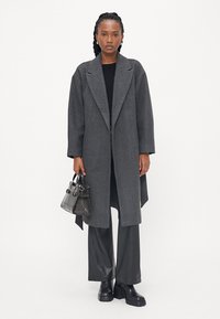 Cappotto grigio di lana con revers a punta, vestibilità ampia, indossato sopra un top nero, abbinato a pantaloni in pelle nera e una borsa nera con texture.