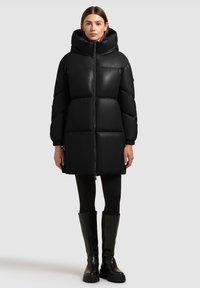 Zwarte donsjas met grote capuchon, ritssluiting aan de voorkant en een zachte textuur. Heeft een doorgestikt ontwerp en elastische manchetten, gecombineerd met zwarte leggings en laarzen.