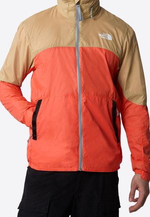 Homme portant une veste North Face à fermeture éclair complète orange et beige avec un pantalon noir, une main dans la poche, debout devant un fond uni.