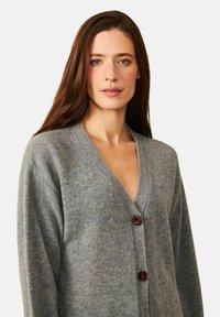 Grå ullkofta med V-ringning och tre bruna knappar fram. Texturen ser mjuk och mysig ut, med en avslappnad passform.